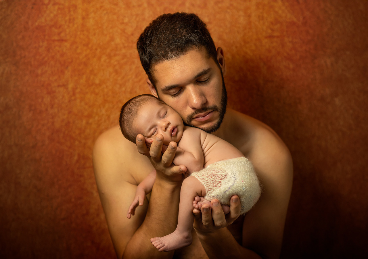 Pai sem camisa segurando bebê recém-nascido no colo em ensaio newborn