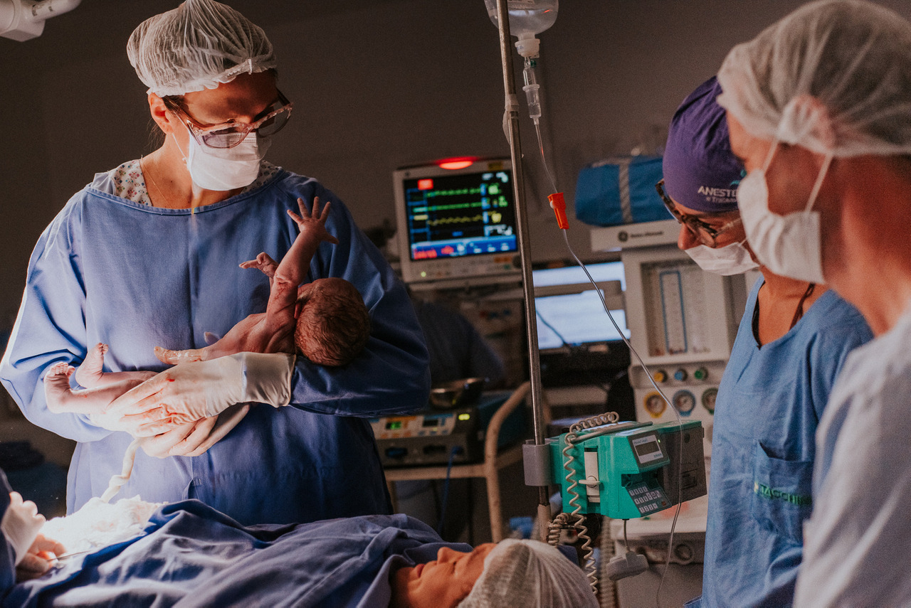 Equipe médica realizando os primeiros cuidados com o recém-nascido após o parto em centro cirúrgico, registro fotográfico de parto com profissionais e equipamentos hospitalares ao fundo