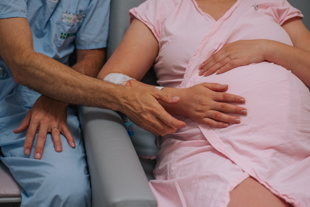 Gestante sentada em ambiente hospitalar com as mãos do parceiro e do profissional de saúde apoiadas sobre a barriga, fotografia documental relacionada ao acompanhamento do parto