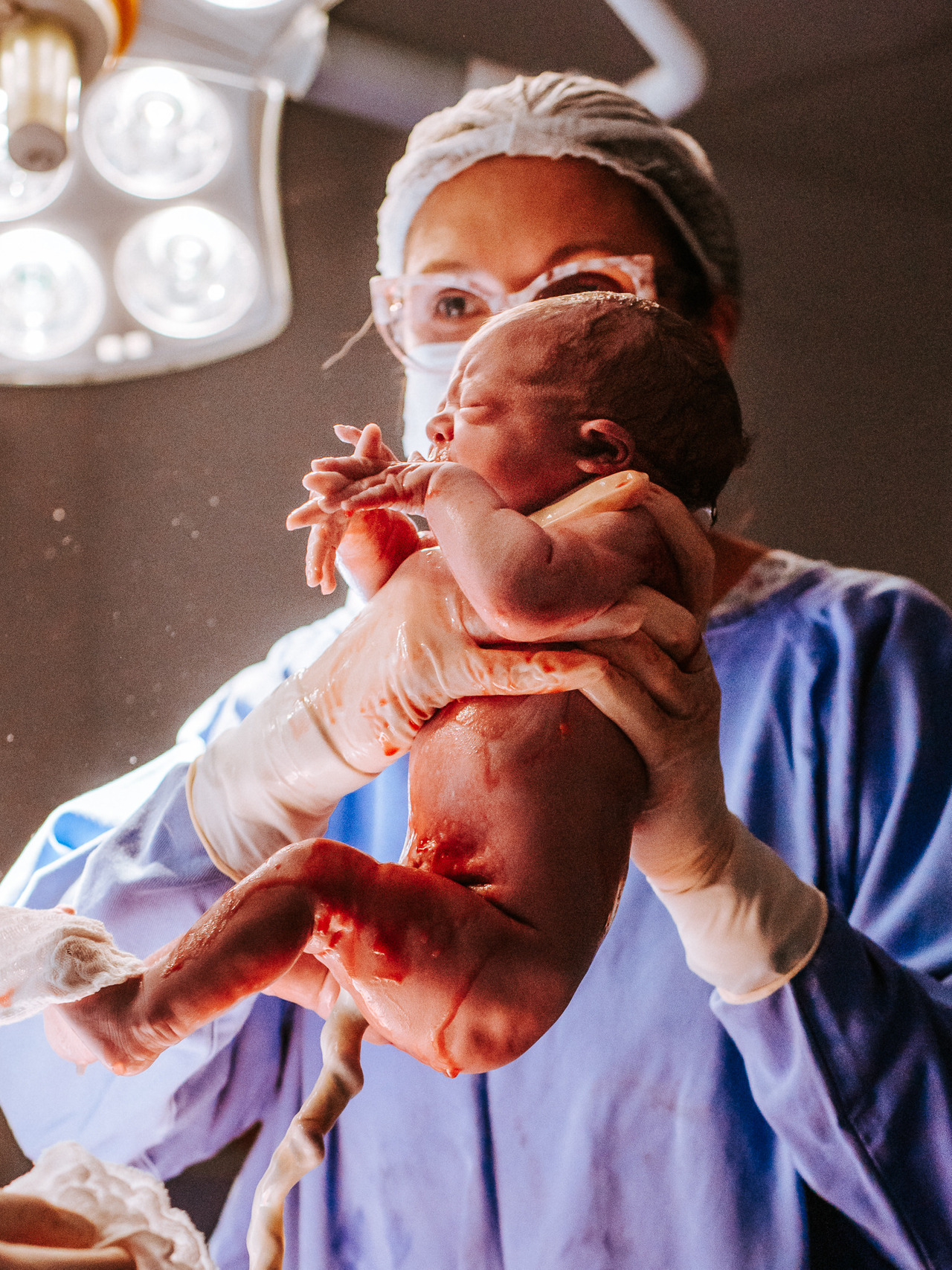 Recém-nascido sendo erguido logo após o nascimento em sala cirúrgica, fotografia de parto registrando os primeiros instantes de vida com equipe médica ao fundo