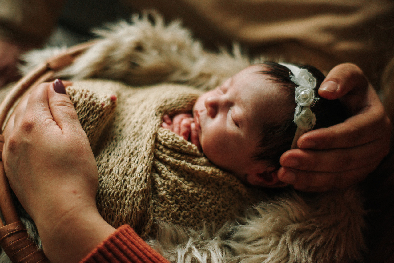 Bebê recém-nascido dormindo com manta durante ensaio newborn, protegido pelas mãos dos pais, representando fotografia newborn consciente e uso responsável de imagens de bebês.