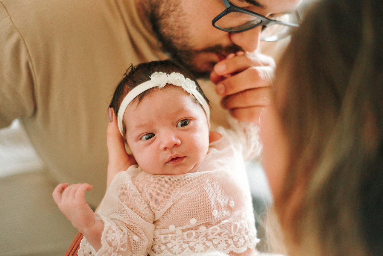 Pais seguram bebê recém-nascido vestido durante ensaio newborn lifestyle, representando uma fotografia de bebê cuidadosa para portfólio profissional.