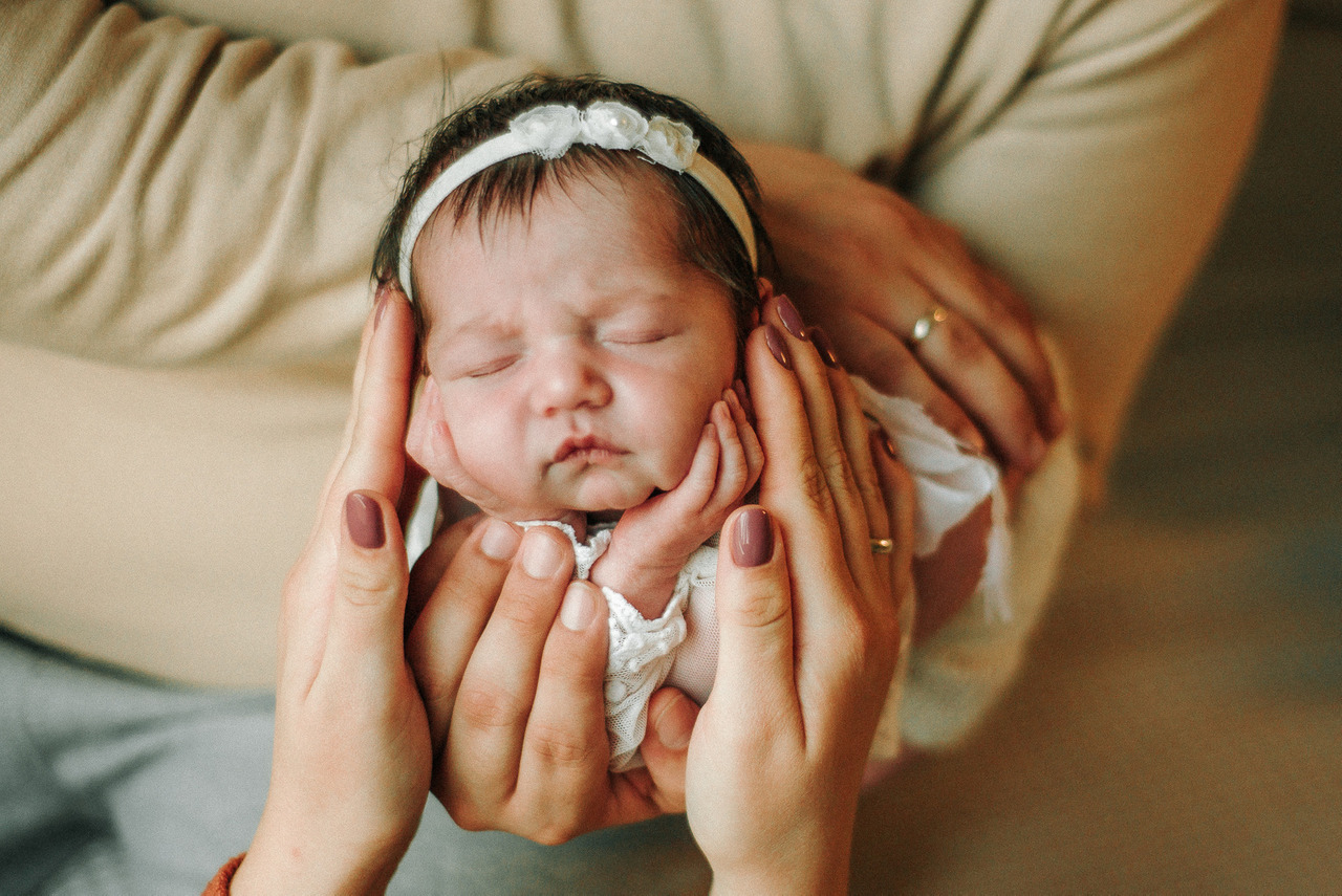 Bebê recém-nascida dormindo com as mãos dos pais ao redor do rosto durante ensaio newborn tradicional com foco em cuidado, vínculo familiar e uso responsável da imagem infantil.