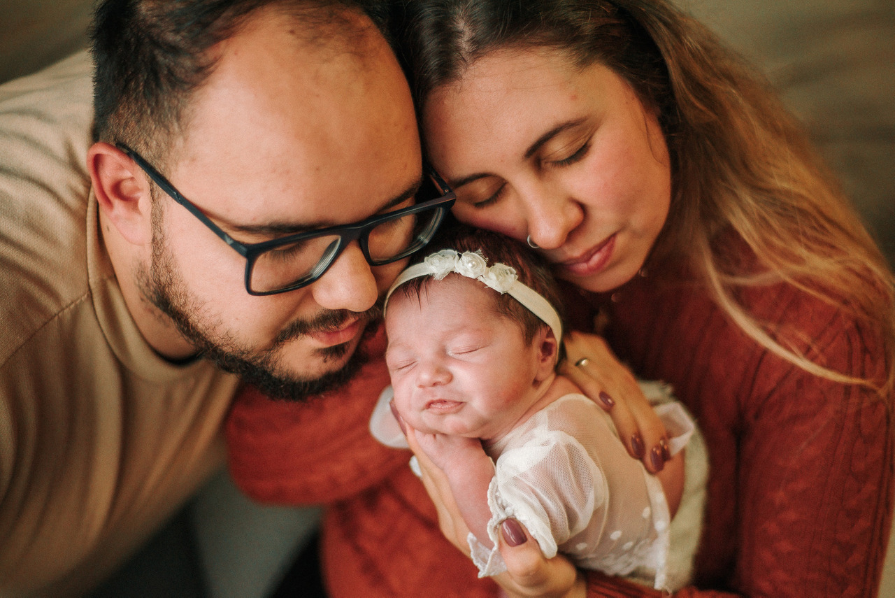 Pais abraçam bebê recém-nascida vestida durante ensaio newborn lifestyle, representando fotografia de bebê com vínculo familiar, cuidado e responsabilidade no uso da imagem infantil.
