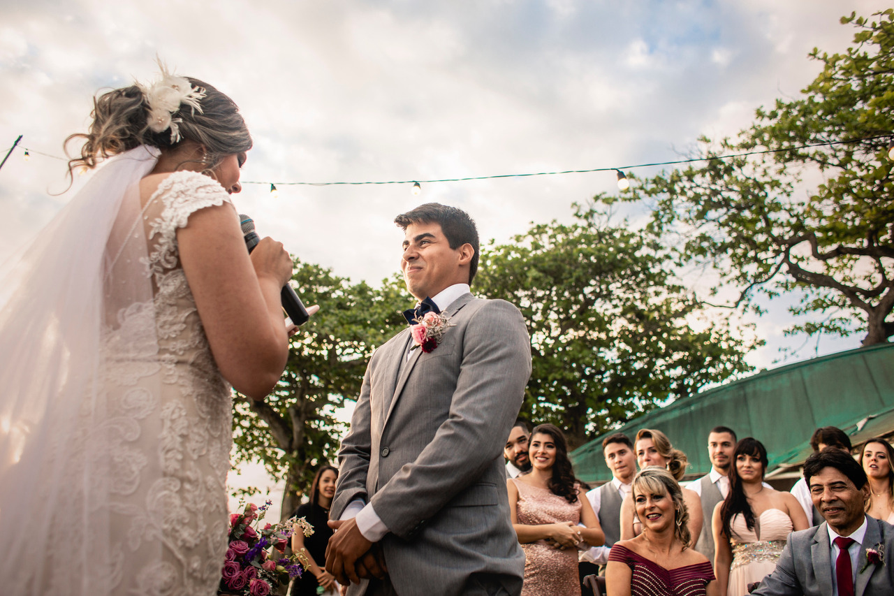 Foto em cores. Noivos um em frente ao outro durante cerimônia de casamento. Convidados ao fundo sorriem. Noiva com microfone na mão e próximo à boca. Noivo com expressão facial de emocionado.