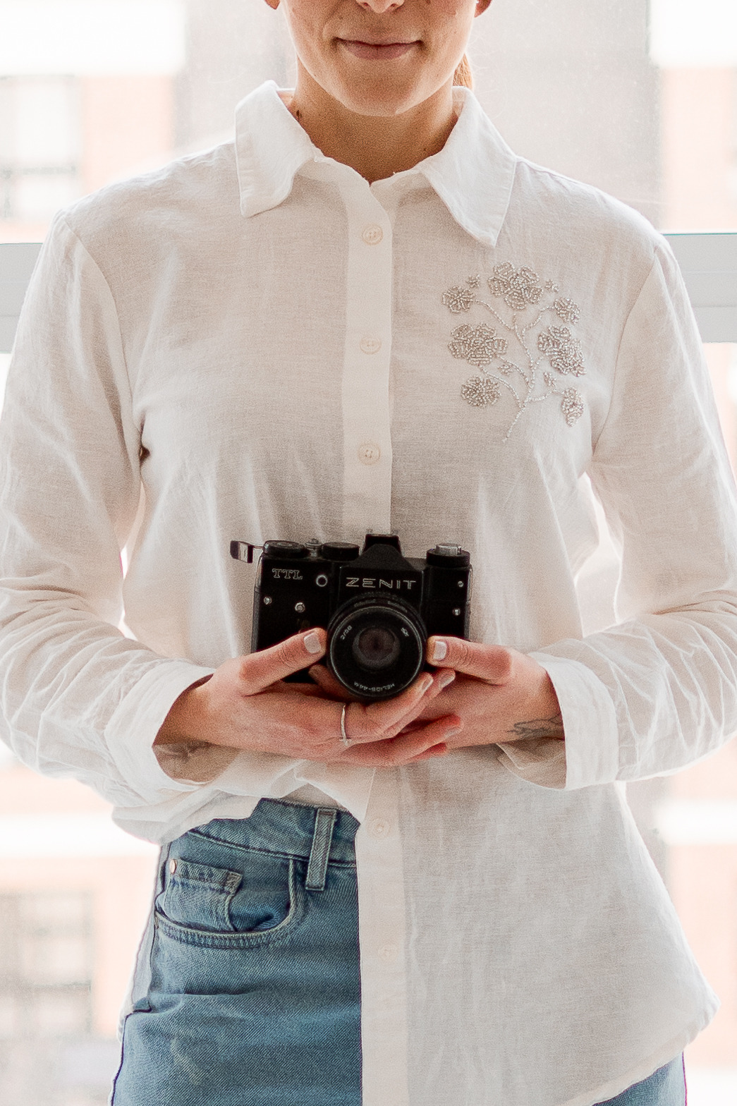 Fotógrafa Flávia, mulher loira e alta, em pé, sorrindo, encostada na parede segurando uma câmera fotográfica na mão. Ao seu lado tem um quadro escrito "a beleza se encontra nos detalhes"