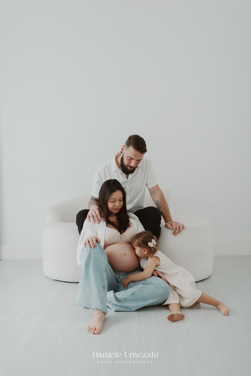 Família em ensaio gestante em casa em Mogi das Cruzes, com mãe, pai e filha sentados juntos em clima afetivo e acolhedor. Fotografia por Dani Umezaki.
