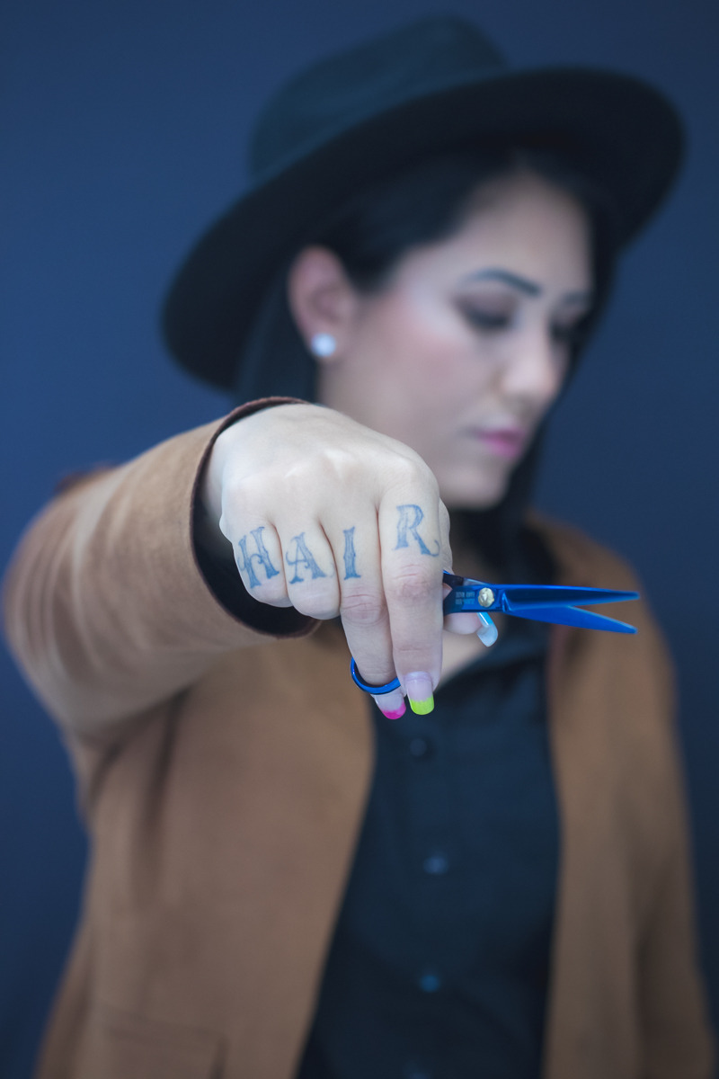 Mulher, cabeleireira usando chapéu e camisa preta, brinco em pérola e blazer em camurça ferrugem. Cabeça levemente inclinada pra baixo, rosto levemente desfocado, segurando uma tesoura à frente do corpo. Nos dedos da mão unhas coloridas e uma tatuagem em cada falangeformando a palavra hair.
