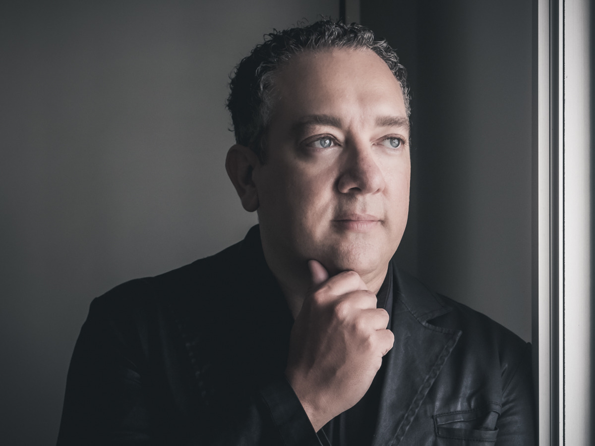 Retrato de um homem, braços cruzados, uma das mãos no queixo. trajando camisa e blazer pretos próximo a uma janela onde sua imagem está refletida 