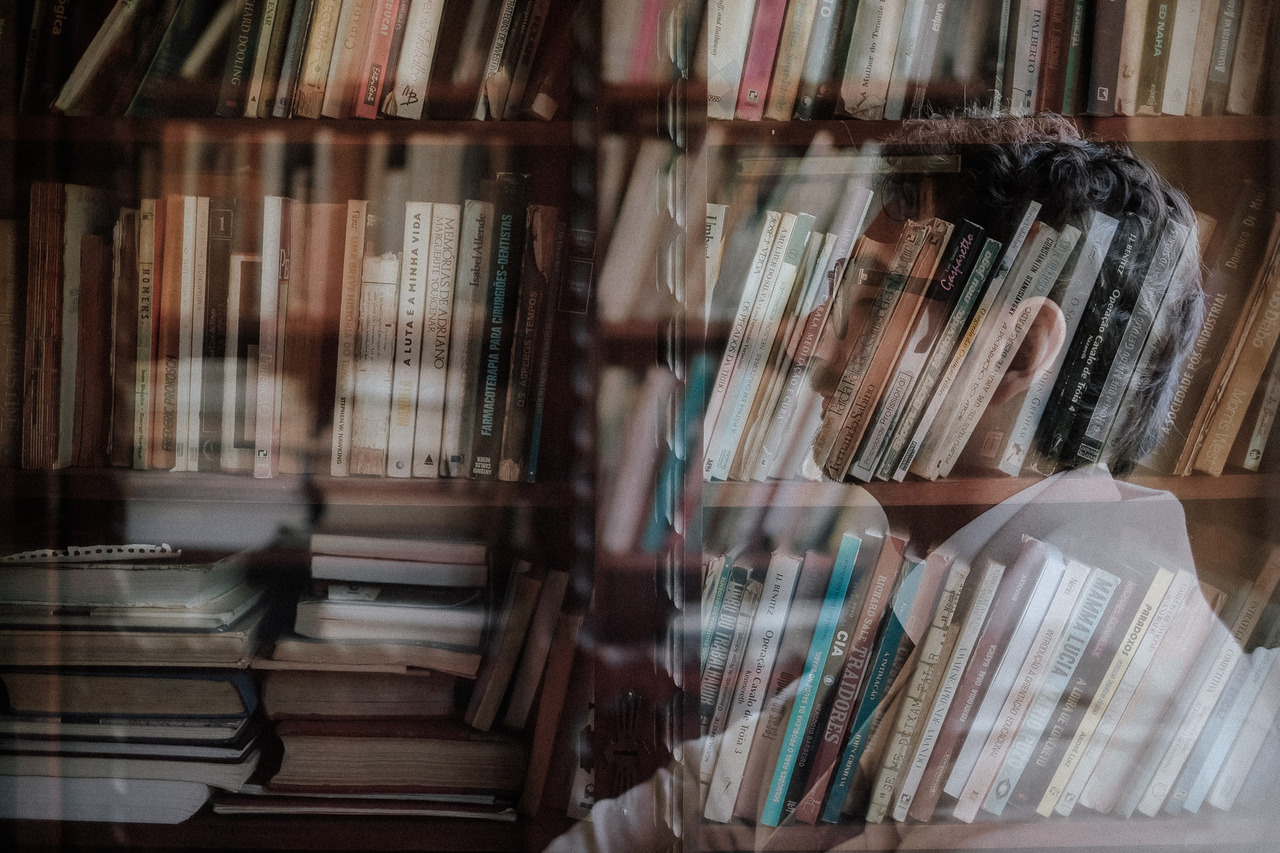 homem de óculos no seu making of antes da cerimônia começar sentado com corpo e rosto virado para a esquerda com uma foto de livros por cima de toda a foto