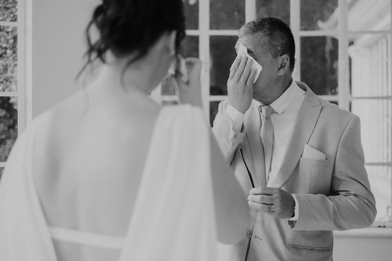 pai se emociona ao ver a filha vestida de noiva. na foto a noiva aparece de costas à esquerda e seu pai aparece enxugando as lágrimas à direita. fotografia em preto e branco.