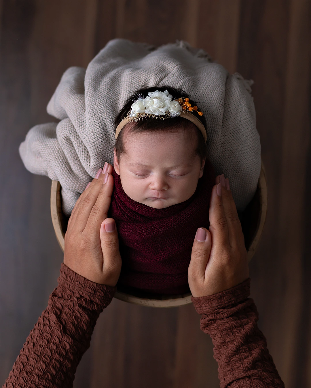 bebê recém-nascido enrolado e dentro de baldinho visto de cima. A foto mostra o rostinho de uma menina que usa uma tiara com flores na cabeça. As mãos da mãe estão em volta dela.