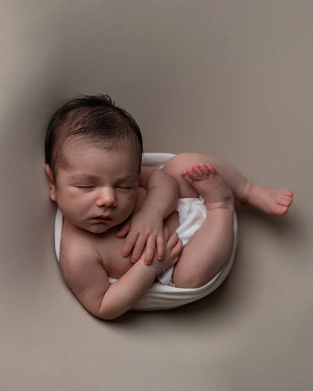 bebê em ensaio Newborn. Ele está deitado com mãos e pés a amostra. Seu rostinho tem destaque e seu corpo está enrolado em um tecido branco.