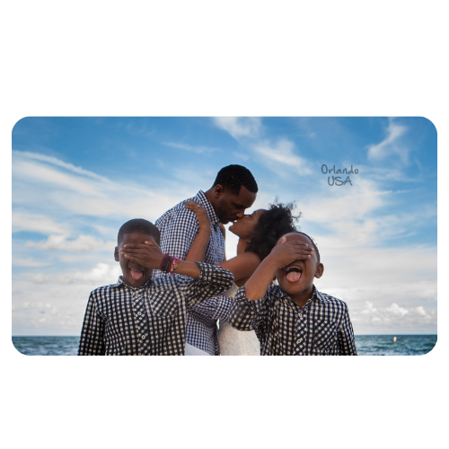 Casal se beija ao fundo em uma praia enquanto os dois filhos estão na frente da foto fechando os olhos e mostrando a lingua