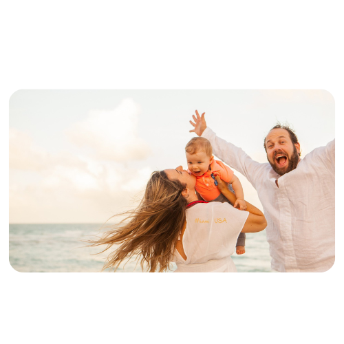 Casal na praia, a mulher segura seu bebê enquanto sorri e o homem está de braços abertos enquanto também sorri