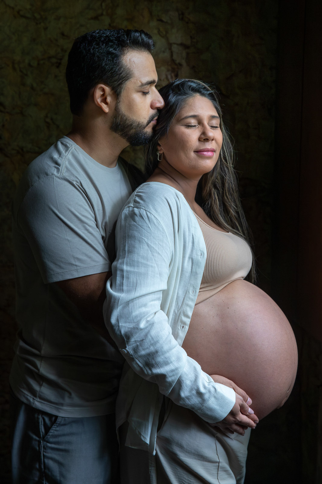 Casal de gestantes abraçados de olhos fechados com luz de janela no Jardim Botânico do Rio de Janeiro