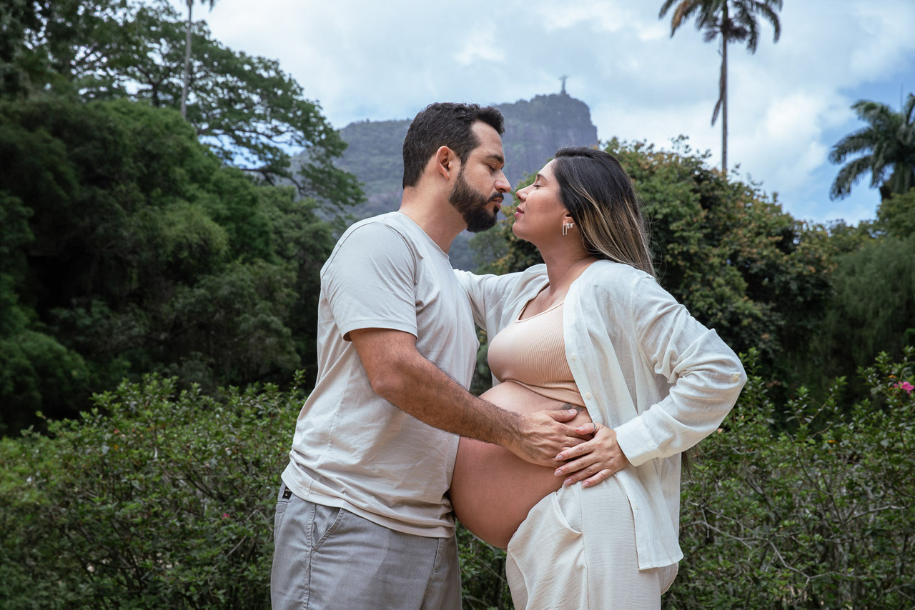 Casal de gestante se abraçando com o Cristo Redentor ao fundo no Jardim Botânico do Rio de Janeiro