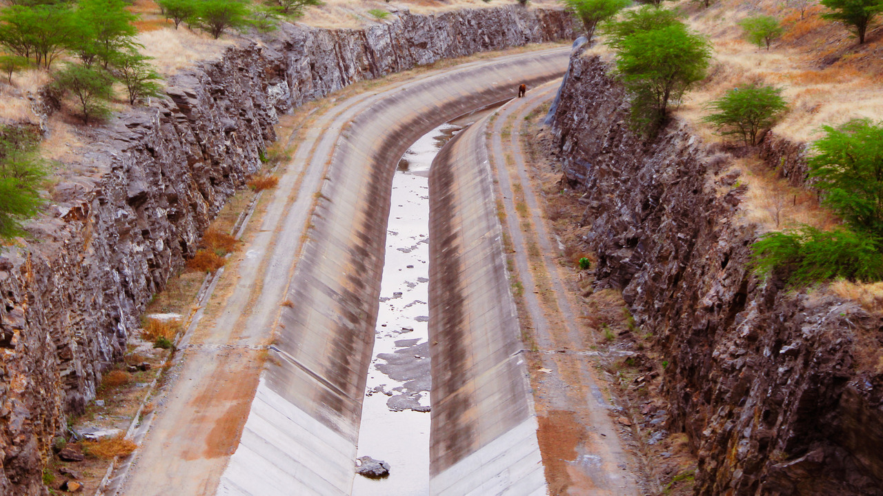 A imagem é uma perspectiva de cima de um canal que está entre barreiras de pedra. Ao fundo o canal faz uma curva para a direita.