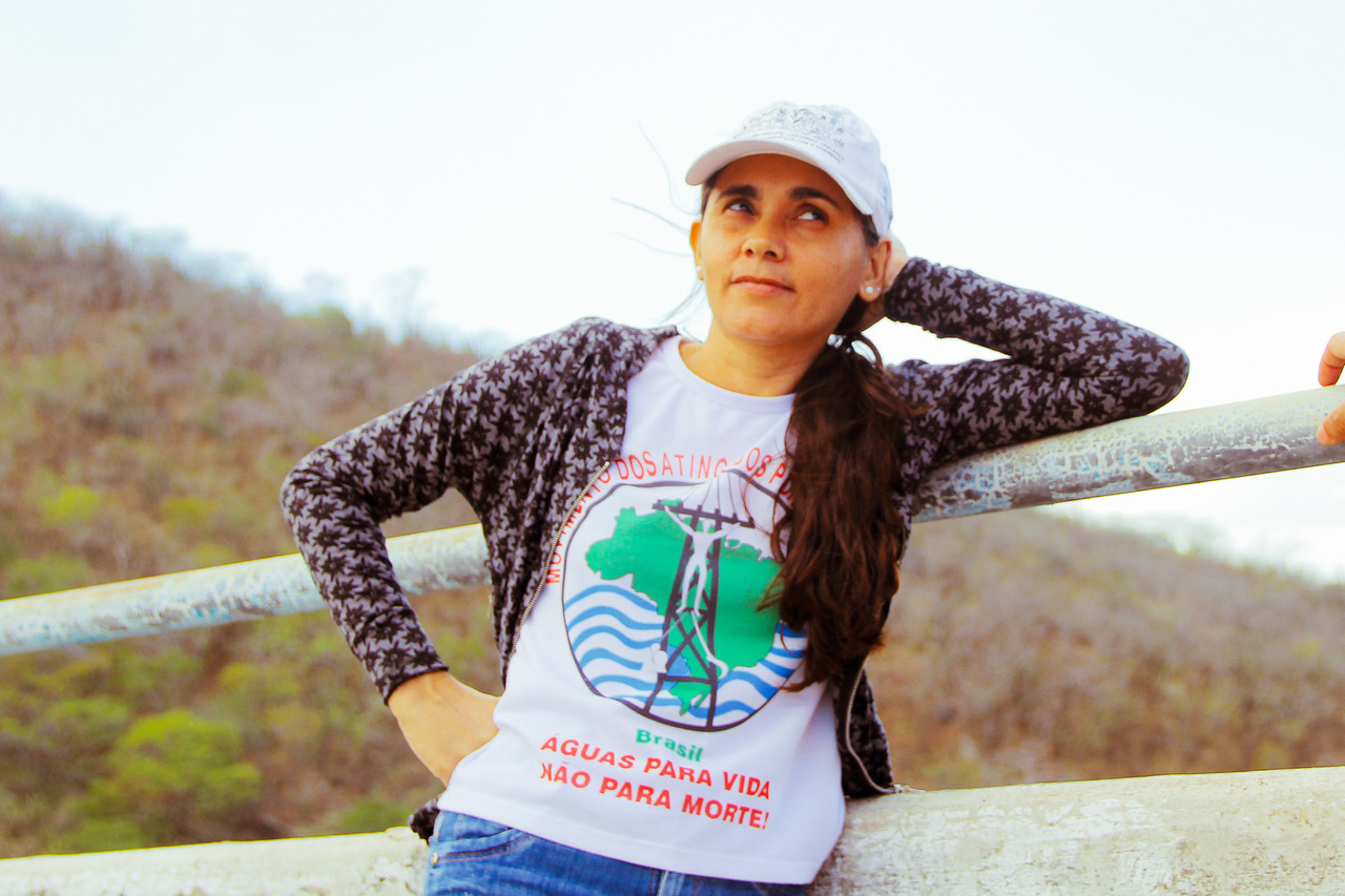 Foto de Aline Araújo, ela é branca, está de cabelos amarrados e com boné branco, veste uma camisa branca com o símbolo do Movimento de Atingidos por Barragem. O símbolo consiste em uma esfera com o mapa do brasil em verde, ondulações em azul, uma torre de energia elétrica e uma pessoa de braços abertos. . Na camisa está os dizeres "águas para a vida e não para a morte"