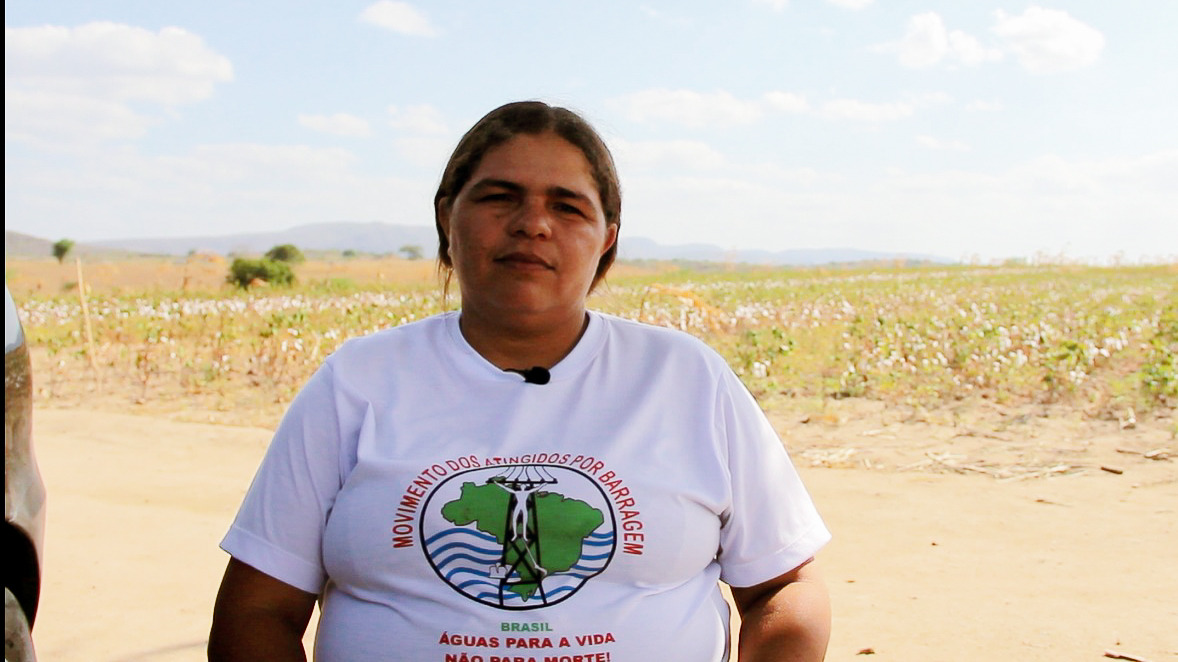 Foto frontal a Joelma, que está sentada, ao fundo um céu azul e vasto plantio de algodão. Joelma está vestida com a camisa do Movimento de atingidos por barragem. Símbola redondo com mapa do brasil em vervende, ondulações na cor azul, torre elétrica em preto e uma pessoa que atravessa todo o mapa
