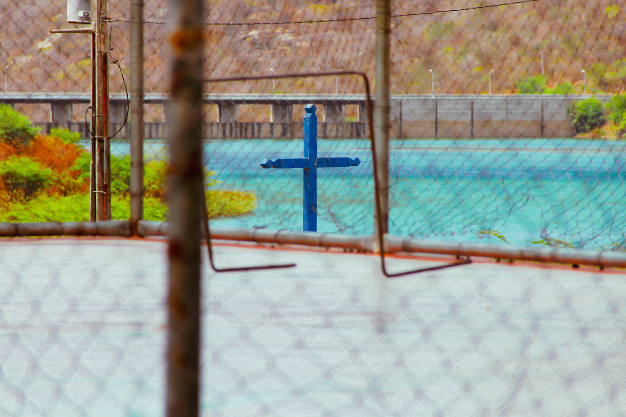 em destaque ao fundo a barragem de Acauã, uma paisagem e muita água. uma cruz está enquadratda em um gol pequeno de futebol