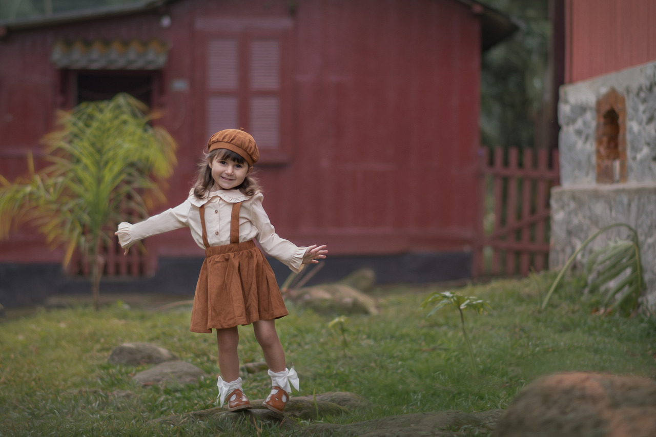 Na imagem, vemos uma menina pequena, com cerca de 3 ou 4 anos, sorrindo enquanto posa ao ar livre. Ela veste um conjunto vintage em tons terrosos, composto por um chapéu e um vestido com suspensórios, combinados com uma camisa de manga longa. Seus sapatinhos marrons e meias brancas com laços completam o visual.   A menina está em pé sobre um gramado, com os braços abertos em uma pose descontraída e alegre, transmitindo leveza e espontaneidade. Ao fundo, há uma casa de madeira pintada de vermelho com detalhes em pedra e cercada por uma vegetação simples, que remete a um ambiente rural ou de vilarejo. A cena parece ter sido capturada em um momento de brincadeira ao ar livre, reforçando a atmosfera acolhedora e lúdica.