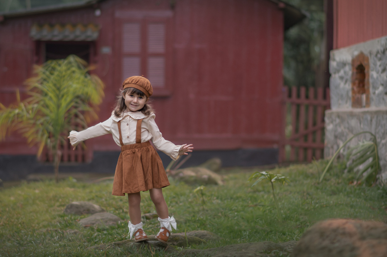 Na imagem, vemos uma menina pequena, com cerca de 3 ou 4 anos, sorrindo enquanto posa ao ar livre. Ela veste um conjunto vintage em tons terrosos, composto por um chapéu e um vestido com suspensórios, combinados com uma camisa de manga longa. Seus sapatinhos marrons e meias brancas com laços completam o visual. A menina está em pé sobre um gramado, com os braços abertos em uma pose descontraída e alegre, transmitindo leveza e espontaneidade. Ao fundo, há uma casa de madeira pintada de vermelho com detalhes em pedra e cercada por uma vegetação simples, que remete a um ambiente rural ou de vilarejo. A cena parece ter sido capturada em um momento de brincadeira ao ar livre, reforçando a atmosfera acolhedora e lúdica.