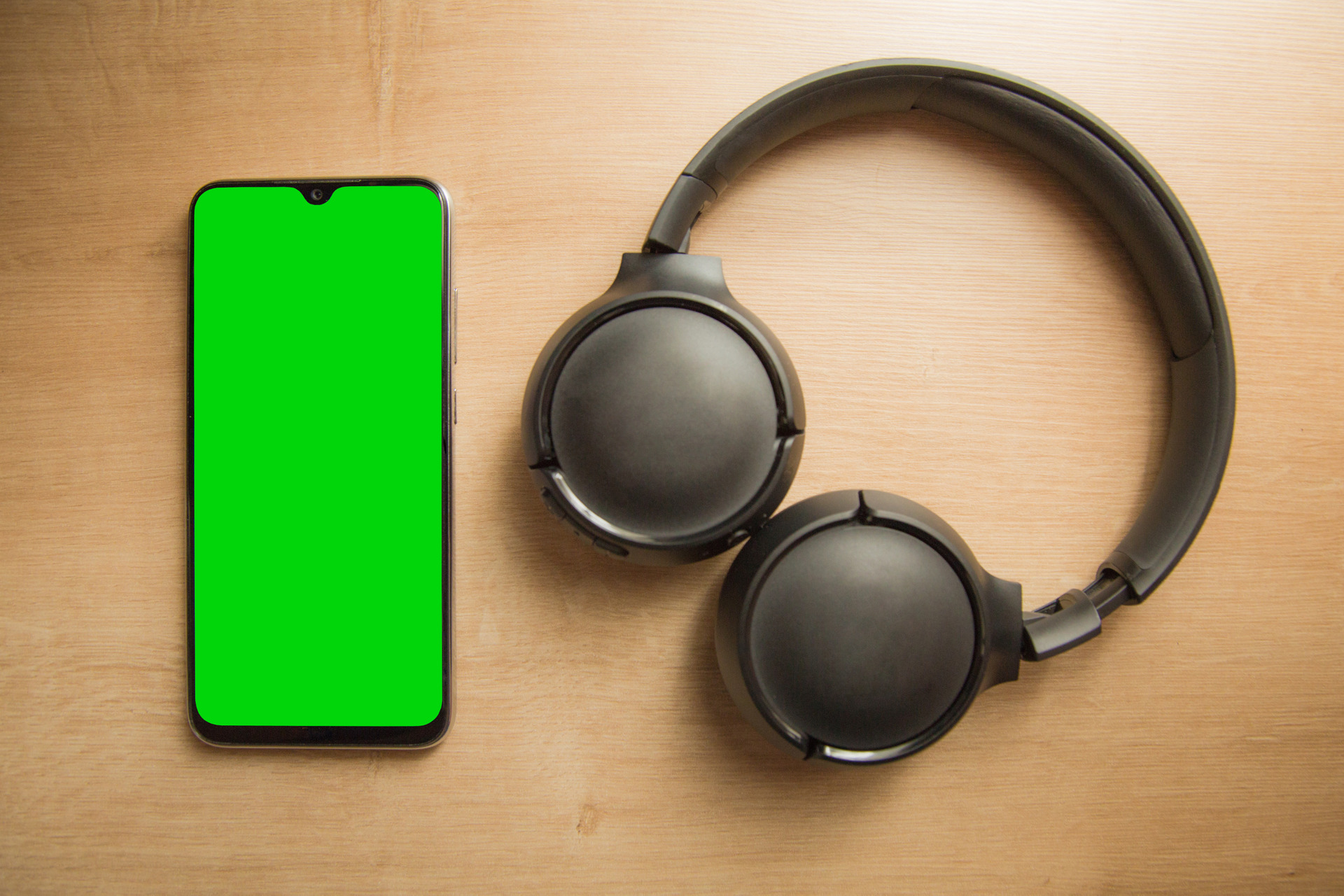 Celular com tela verde (chroma key) e um fone headset sem fio preto sobre uma mesa de madeira, ideal para mockups e projetos de tecnologia ou comunicação.