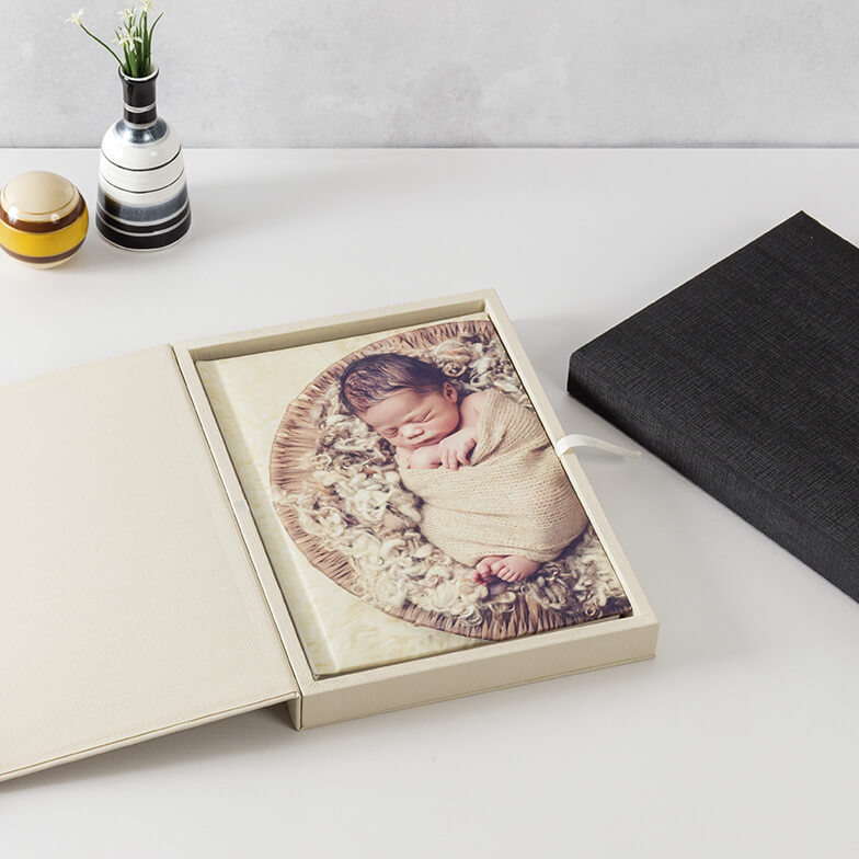 Estojo de álbum profissional na cor marfim, aberto para exibir um álbum com uma foto de newborn na capa. Ao lado, um estojo preto fechado, evidenciando as opções de cores e o design elegante.