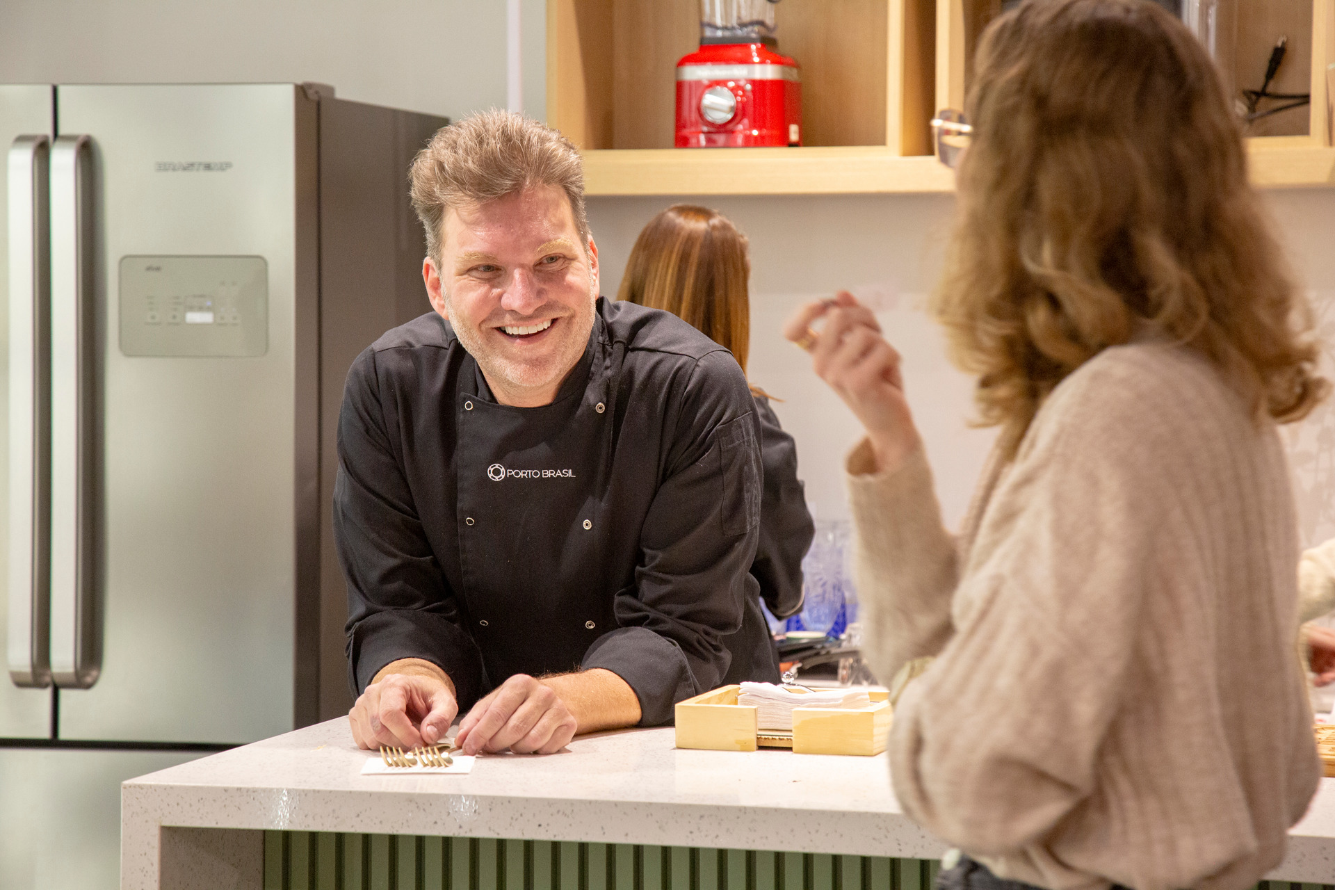 Chef renomado conversando e interagindo com uma participante durante um workshop culinário, demonstrando a troca de conhecimento e a experiência prática.