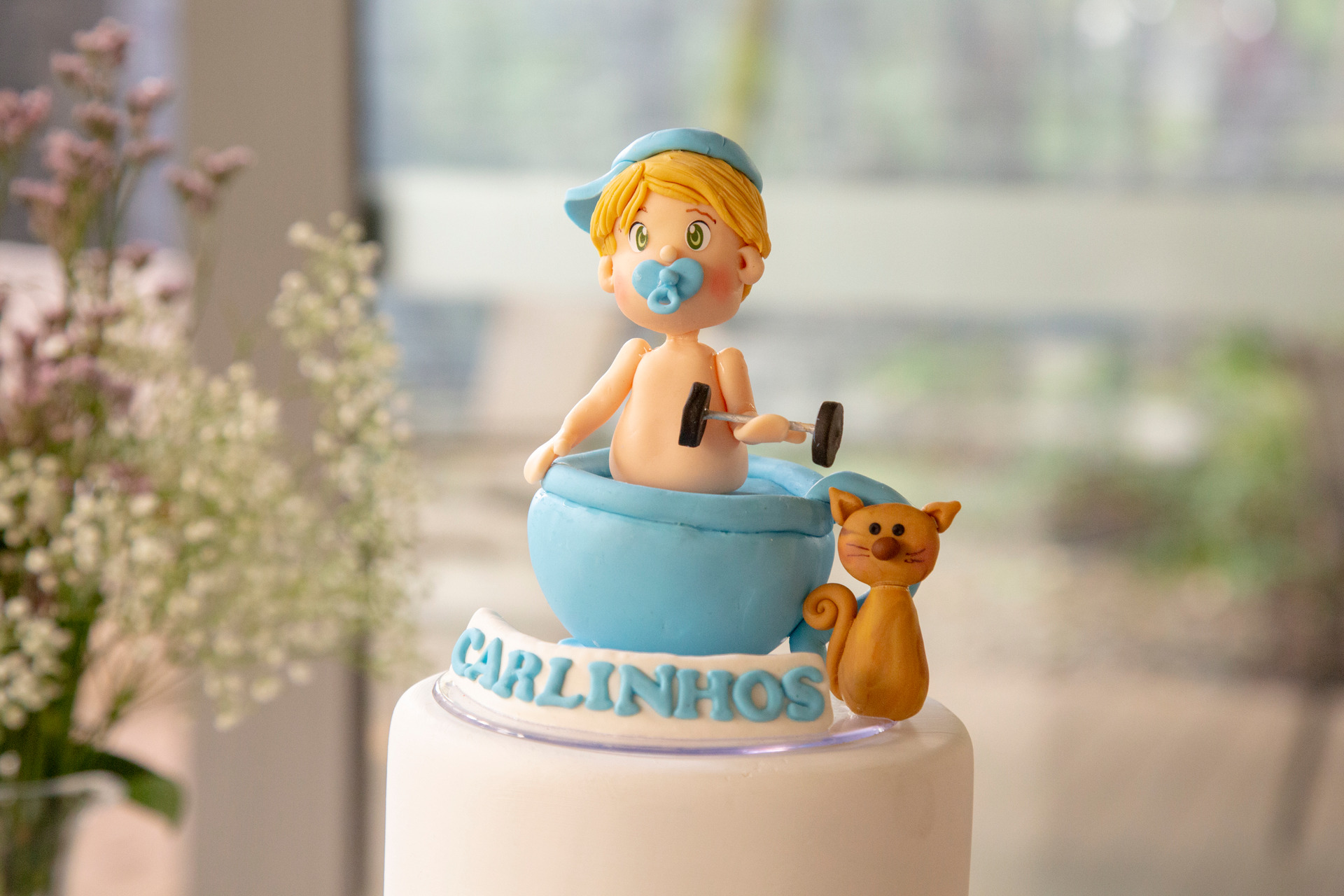 Plano detalhe da decoração do topo de um bolo de chá de bebê, com um boneco de bebê carregando um peso ao lado de um gatinho laranja e o nome 'Carlinhos', mostrando a personalização do evento.