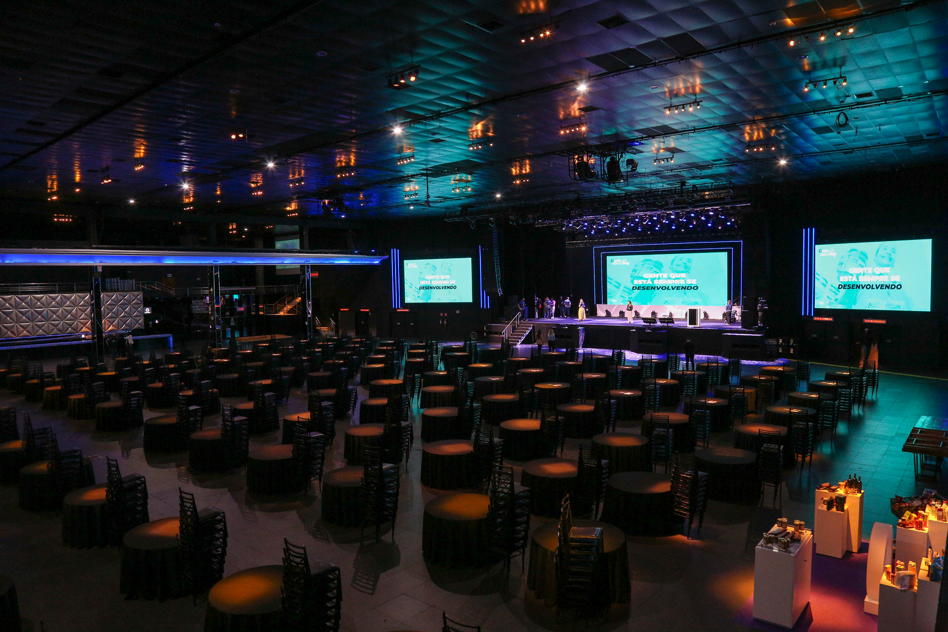 Plano aberto de um grande evento com mesas dispostas para mais de mil pessoas, com um palco e telão ao fundo, mostrando a capacidade de cobertura fotográfica de grandes formatos.