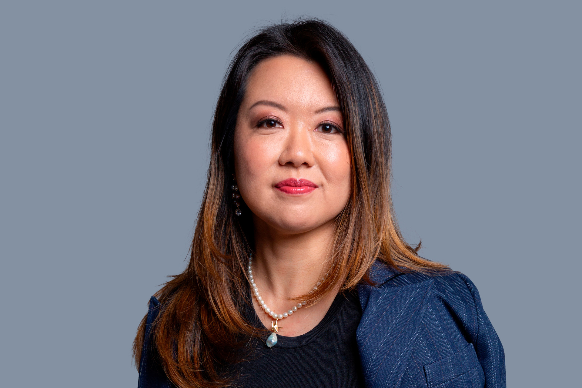 Retrato corporativo de mulher japonesa sentada em pose elegante com blazer azul marinho com listrar brancas finas apoiado nos ombros e colar, com olhar de confiança e amistoso