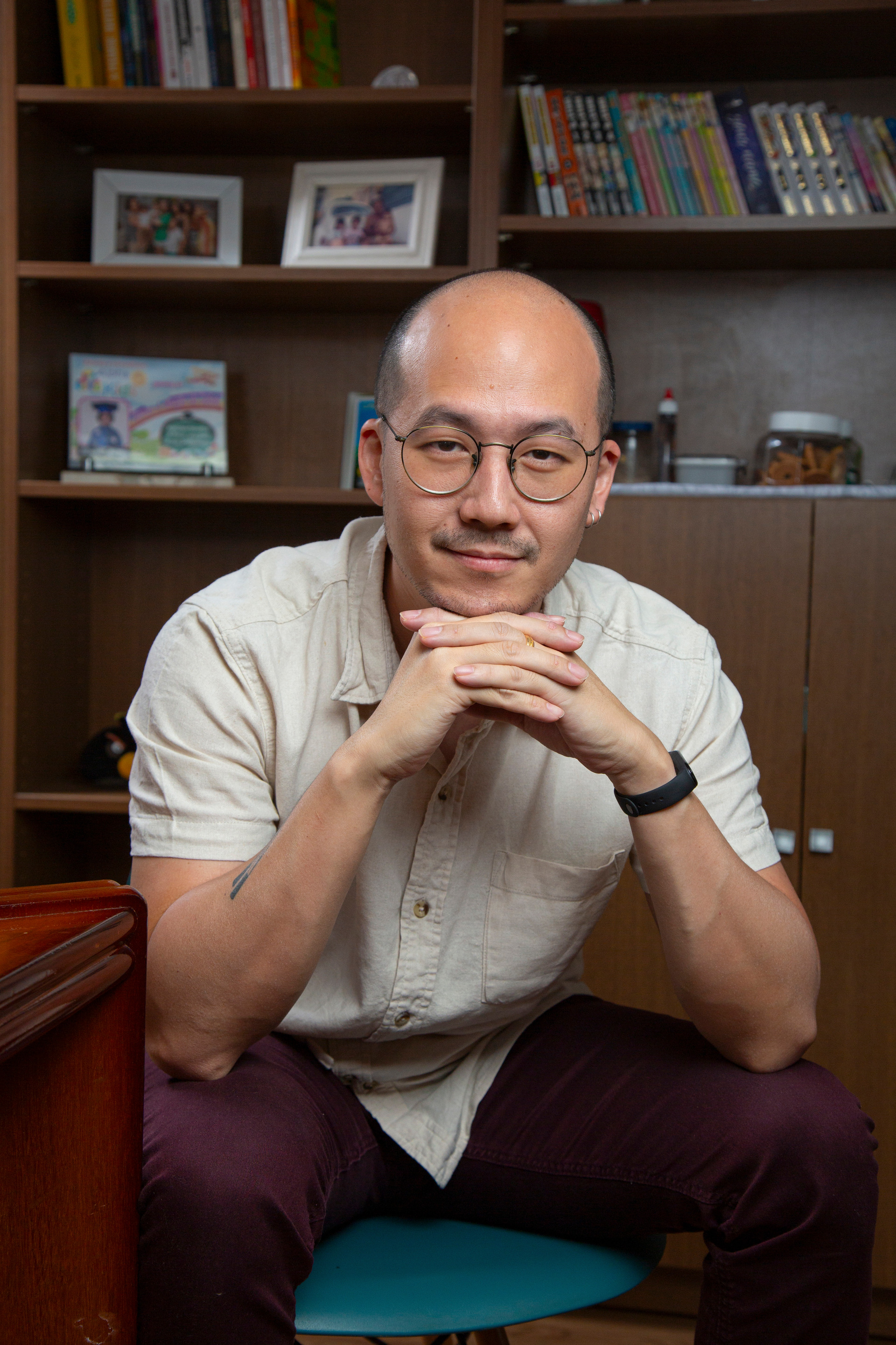 Fotógrafo Bruno Arita sentado, olhando para a câmera com um sorriso amigável, os dedos entrelaçados e o queixo apoiado nas mãos, transmitindo uma postura acessível e confiante.