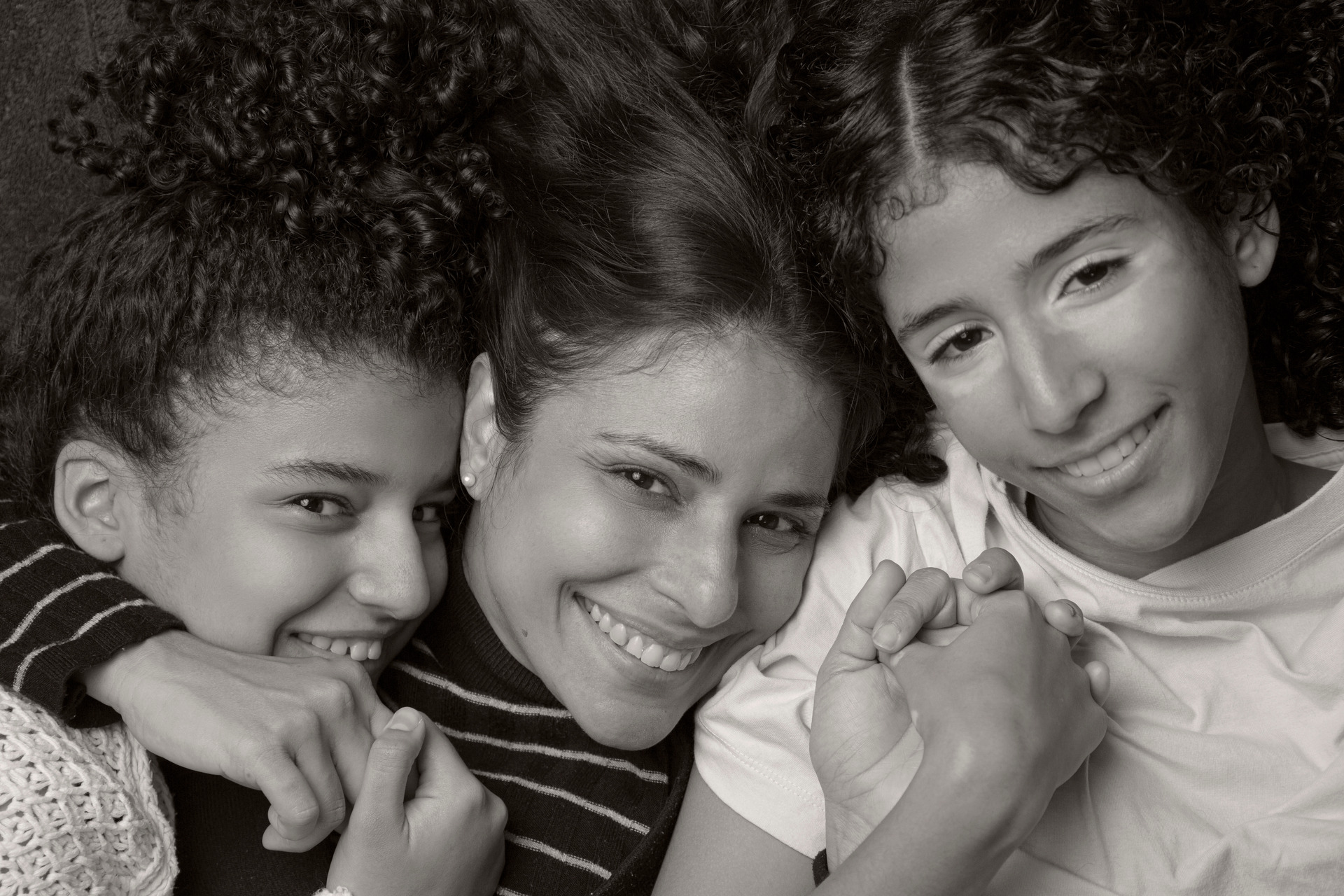 Fotografia de família em preto e branco: mãe sorrindo no centro, com uma filha sorrindo e olhando com amor de cada lado, as mãos da mãe segurando as das filhas, simbolizando conexão e afeto.