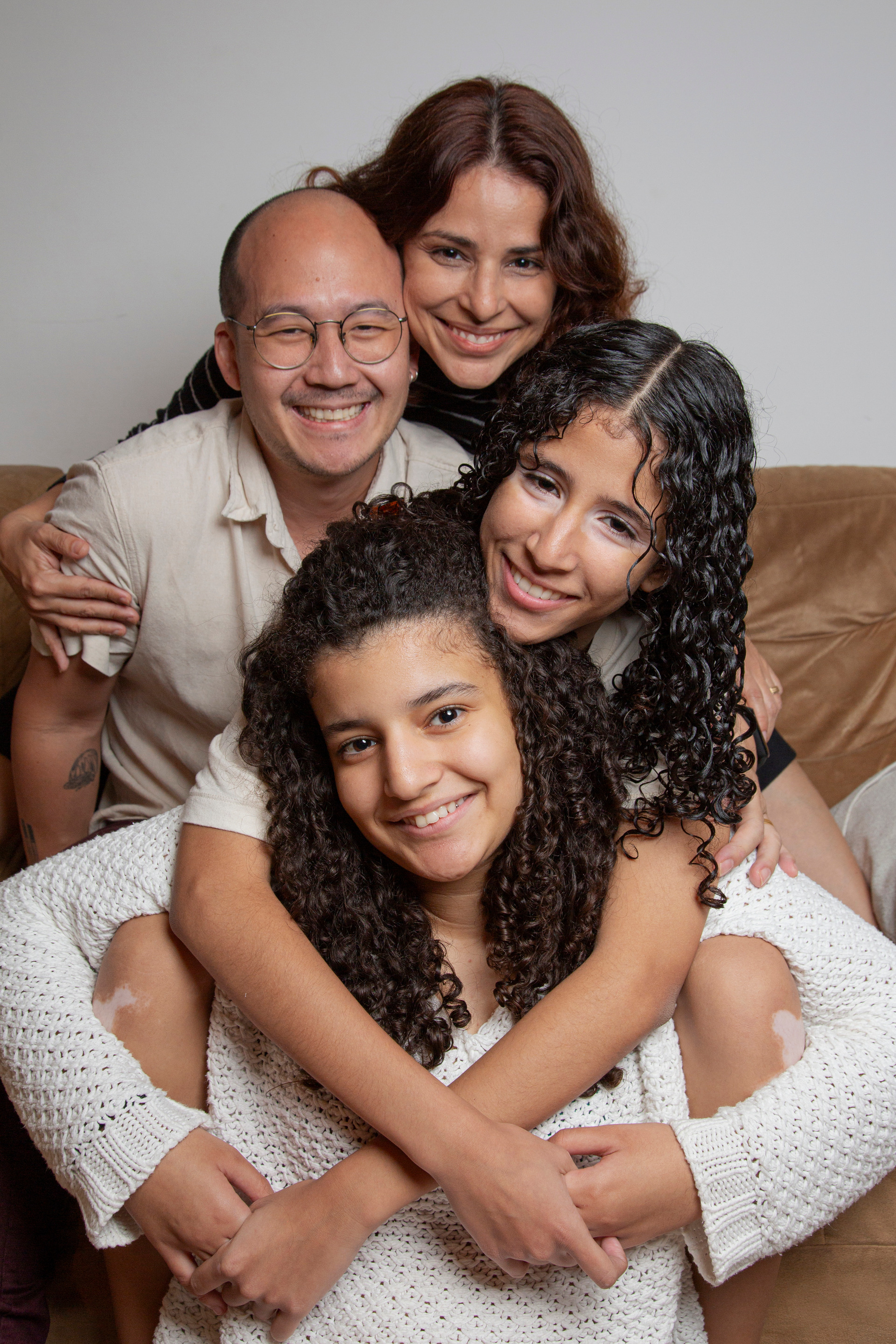 Fotografia de família vertical em camadas: filha sorrindo apoiada na irmã que a abraça, pai atrás da filha que abraça, e mãe apoiada no marido rosto com rosto, todos sorrindo em um momento de alegria e união familiar.