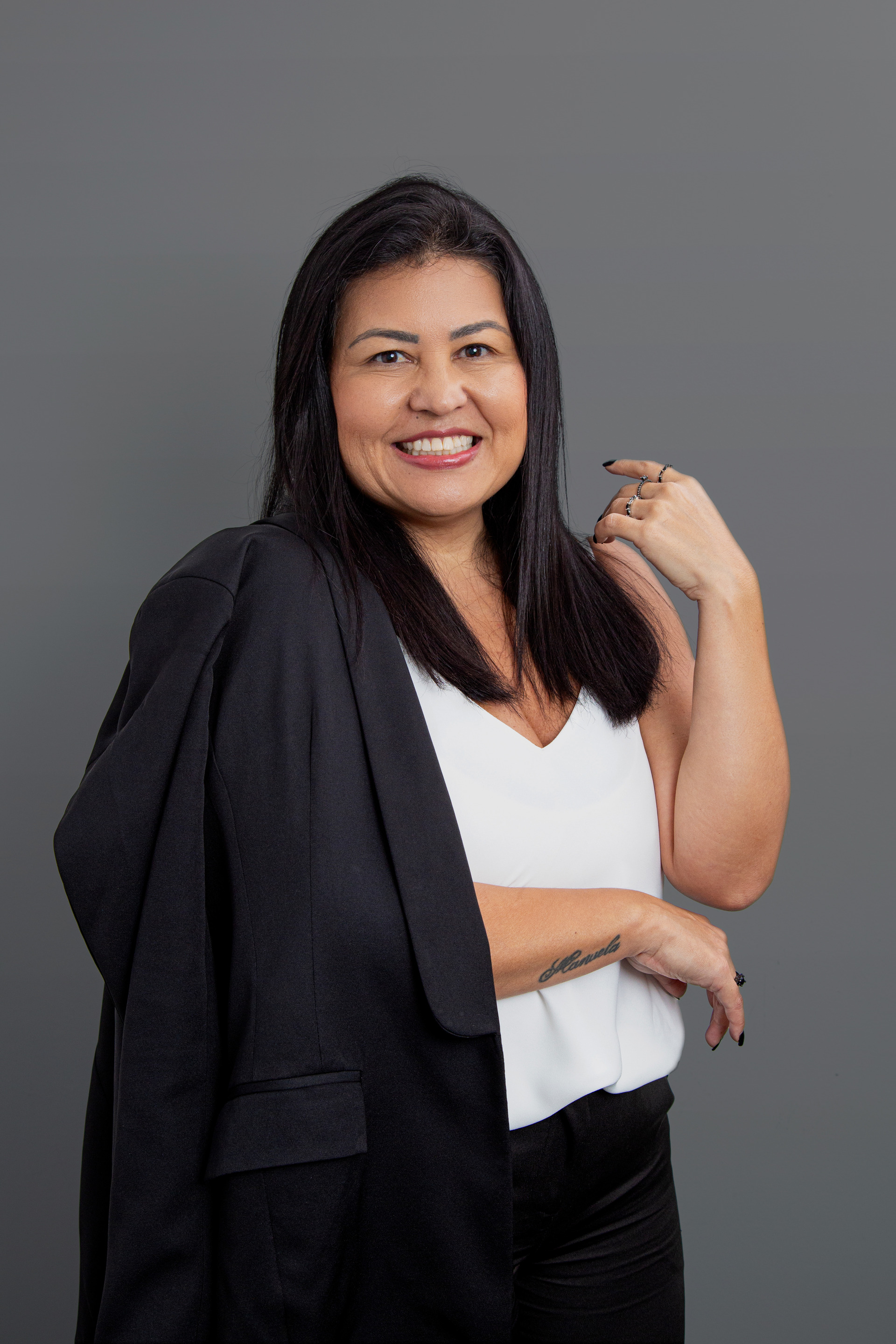Retrato corporativo de uma mulher com traços orientais, na faixa dos 40 anos, cabelo preto, vestindo blusa branca e calça preta social, com um terno preto apoiado em um ombro, em fundo neutro, transmitindo um estilo profissional e moderno.