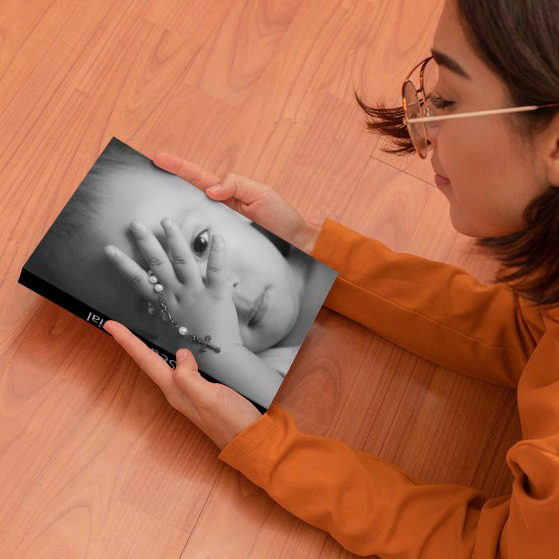 Mulher de óculos observa a capa de um álbum fotográfico fechado, que exibe um retrato em preto e branco de um bebê segurando um terço, transmitindo uma imagem de delicadeza e fé.