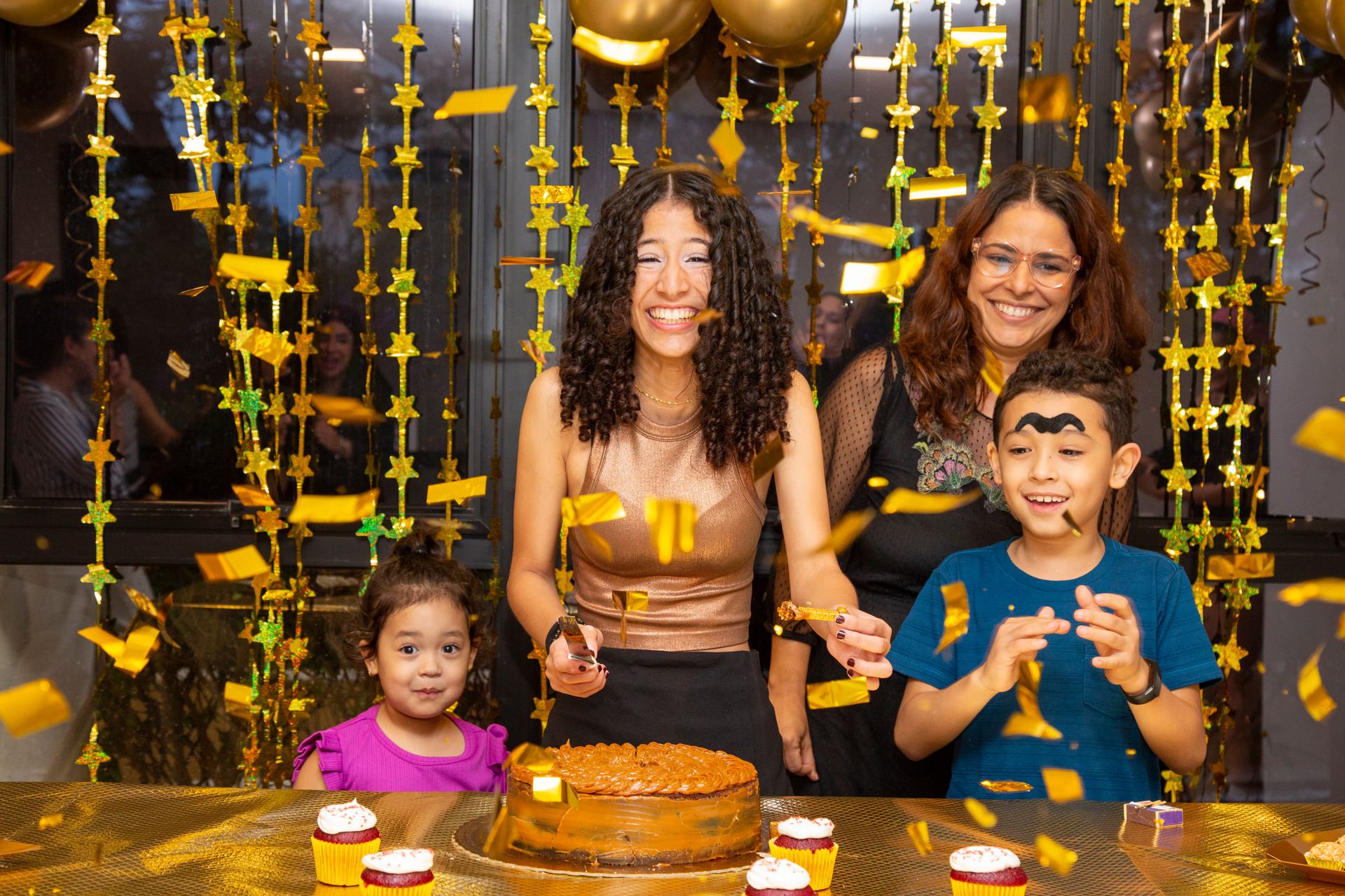 Parabéns de aniversário com explosão de confete, aniversariante ao centro sorrindo surpresa, bolo de churros na mesa dourada, duas crianças de cada lado da aniversariante e uma mulher ao fundo em festa de aniversário em São Paulo