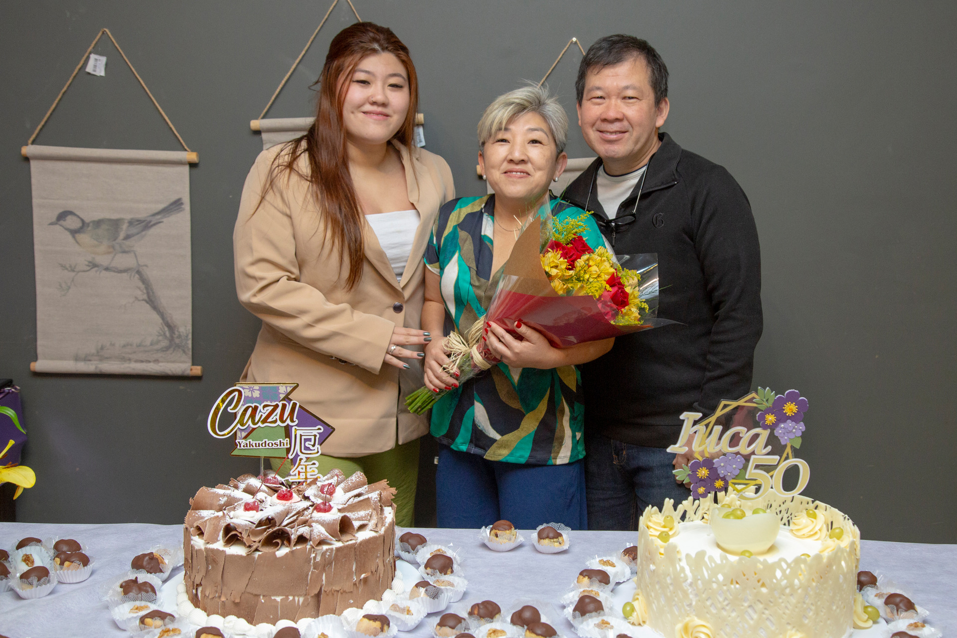 Foto posada de uma mãe aniversariante em São Paulo, sorrindo ao lado da filha adulta e do marido, em uma mesa ricamente decorada com bolos e doces, celebrando seu aniversário adulto em São Paulo e foto de Bruno Arita