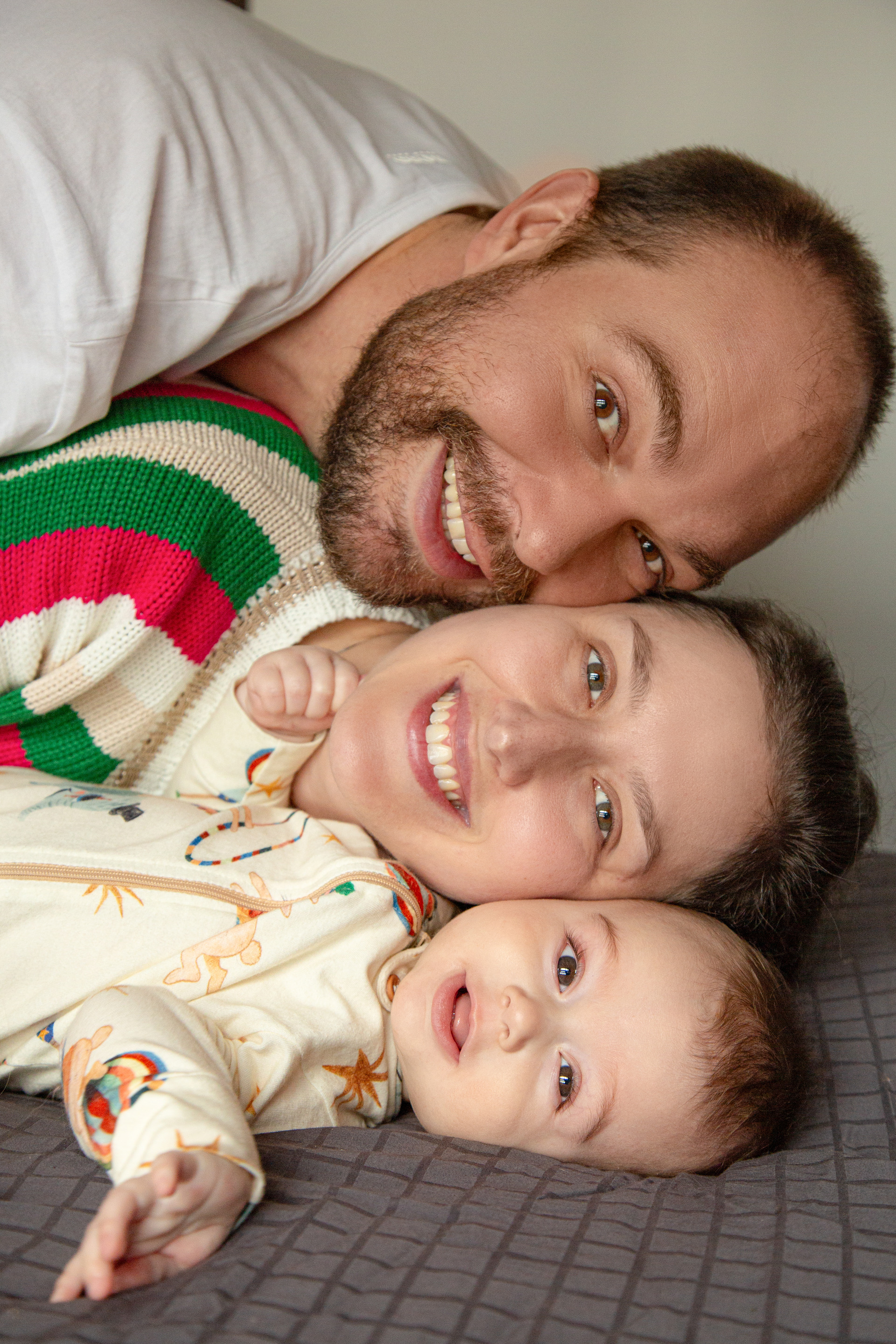 Retrato de família vertical com rostos empilhados na cama: bebê de 6 meses embaixo, mãe no meio e pai por cima, todos sorrindo com expressões de alegria e amor, transmitindo união e carinho no ambiente familiar.