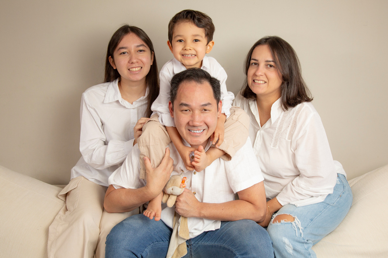 Retrato afetivo de uma família de quatro pessoas com ascendência asiática em um ambiente de estúdio com fundo neutro e iluminação suave. Ao centro, um homem sorri enquanto carrega um menino pequeno nos ombros. À esquerda, uma jovem adolescente e, à direita, uma mulher adulta, ambos sorrindo para a câmera em uma pose acolhedora. Todos vestem camisas brancas coordenadas e calças jeans, transmitindo uma sensação de união, leveza e conexão genuína. Fotografia de Bruno Arita.
