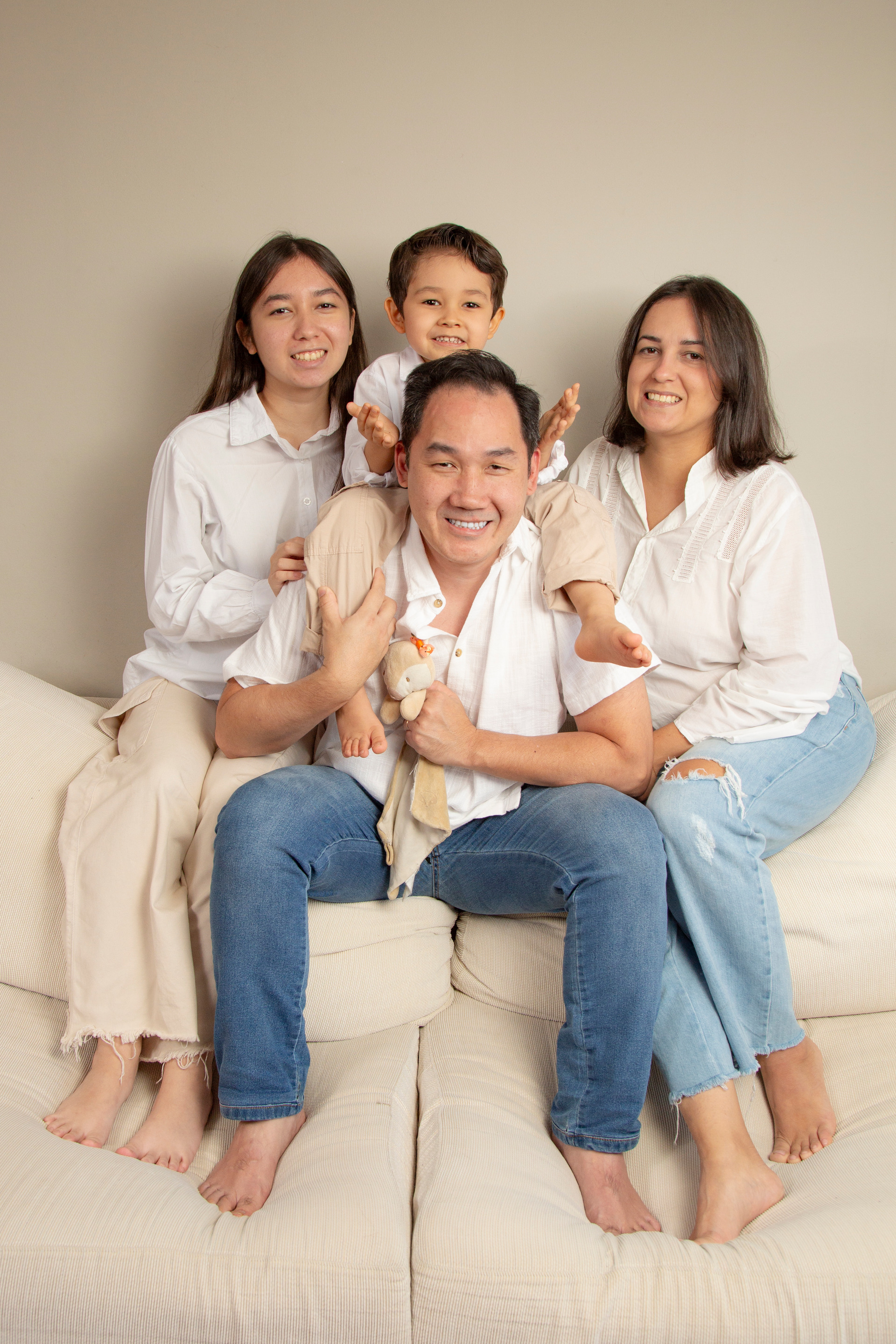 Filha adolescente, filho de 5 anos sentado de cavalinho no pescoço do pai, e mãe (todos de camisa branca/creme e jeans), sentados juntos no encosto alto do sofá, em momento de diversão. (Ensaio de Família, São Paulo, Fotógrafo Bruno Arita)
