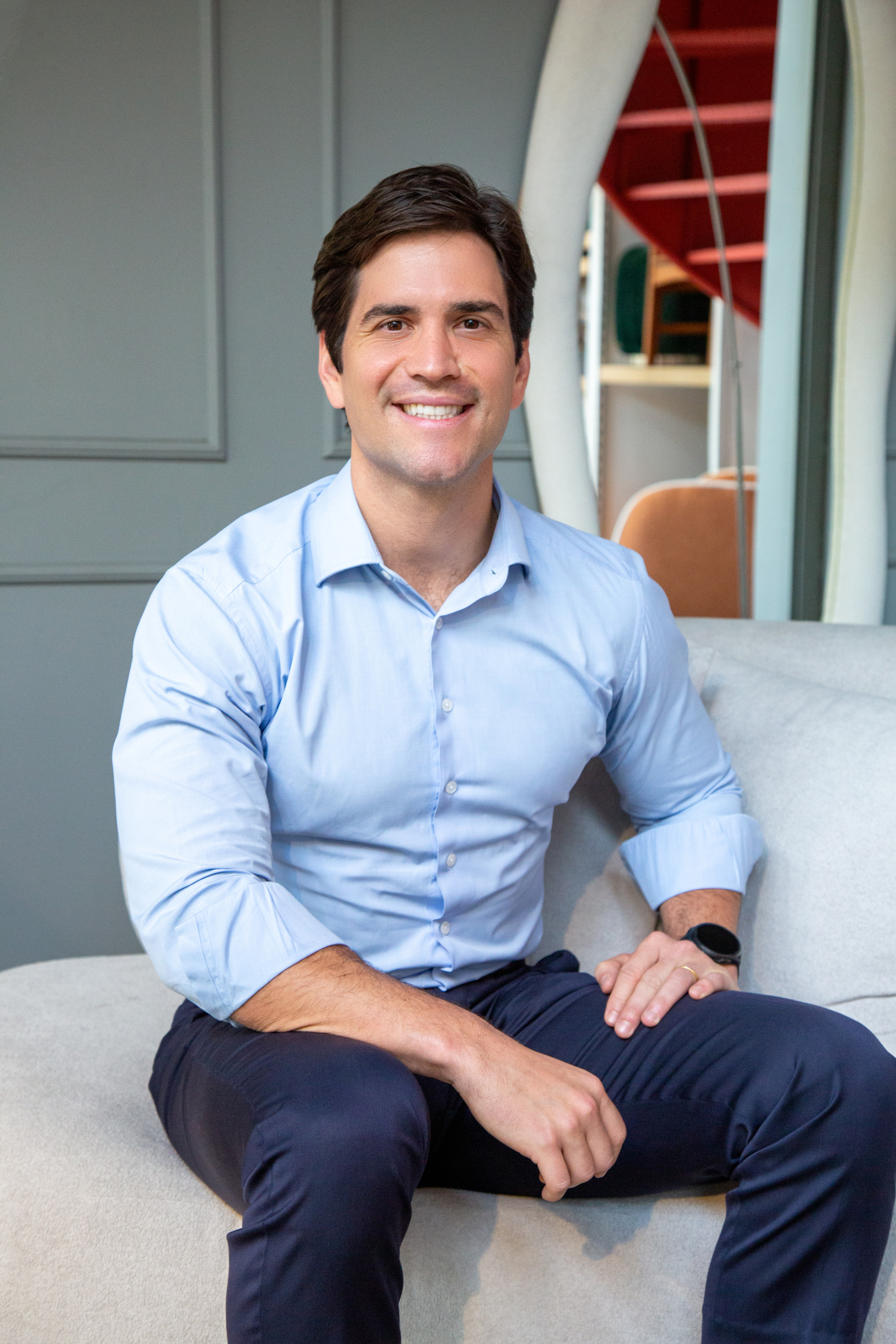 Retrato corporativo de um homem branco, aproximadamente 45 anos, vestindo traje social com camisa azul clara, calça azul marinho, sentado com relógio no pulso com ar de profissionalismo e sorriso em ambiente refinado