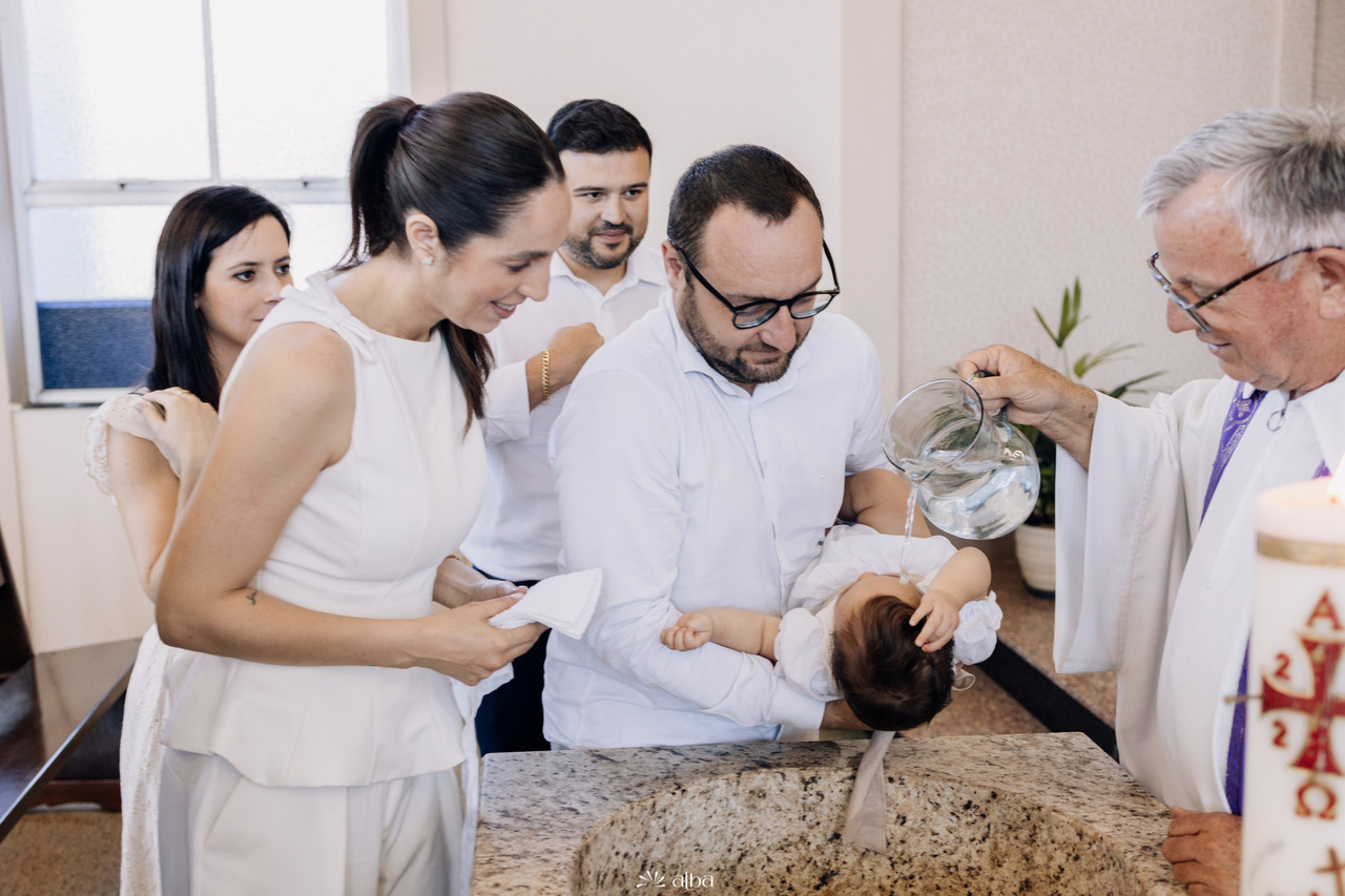 Batizado da Amália na Paróquia São Cristóvão, em Lajeado, com o momento do derramamento da água benta realizado pelo padre enquanto os pais acompanham emocionados. Foto por Alba Estúdio.