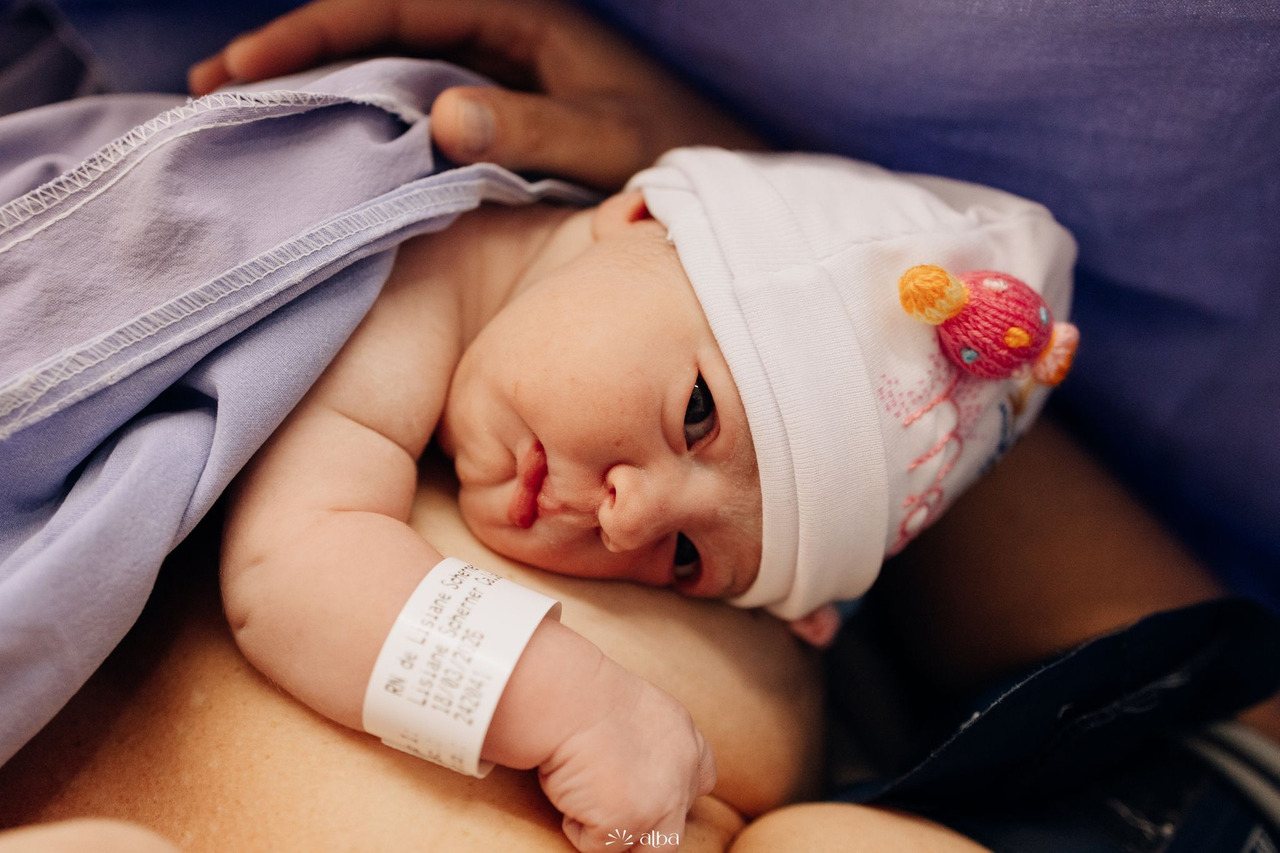 Recém-nascido deitado no colo da mãe, envolto em manta lilás e usando touca branca, em um registro delicado dos primeiros instantes após o nascimento, fotografado pelo Alba Estúdio.  Lia