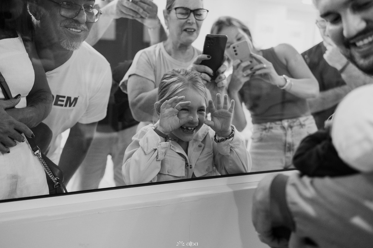 Família reunida do outro lado do vidro da maternidade, observando o recém-nascido com sorrisos e emoção, em um registro em preto e branco feito pelo Alba Estúdio.