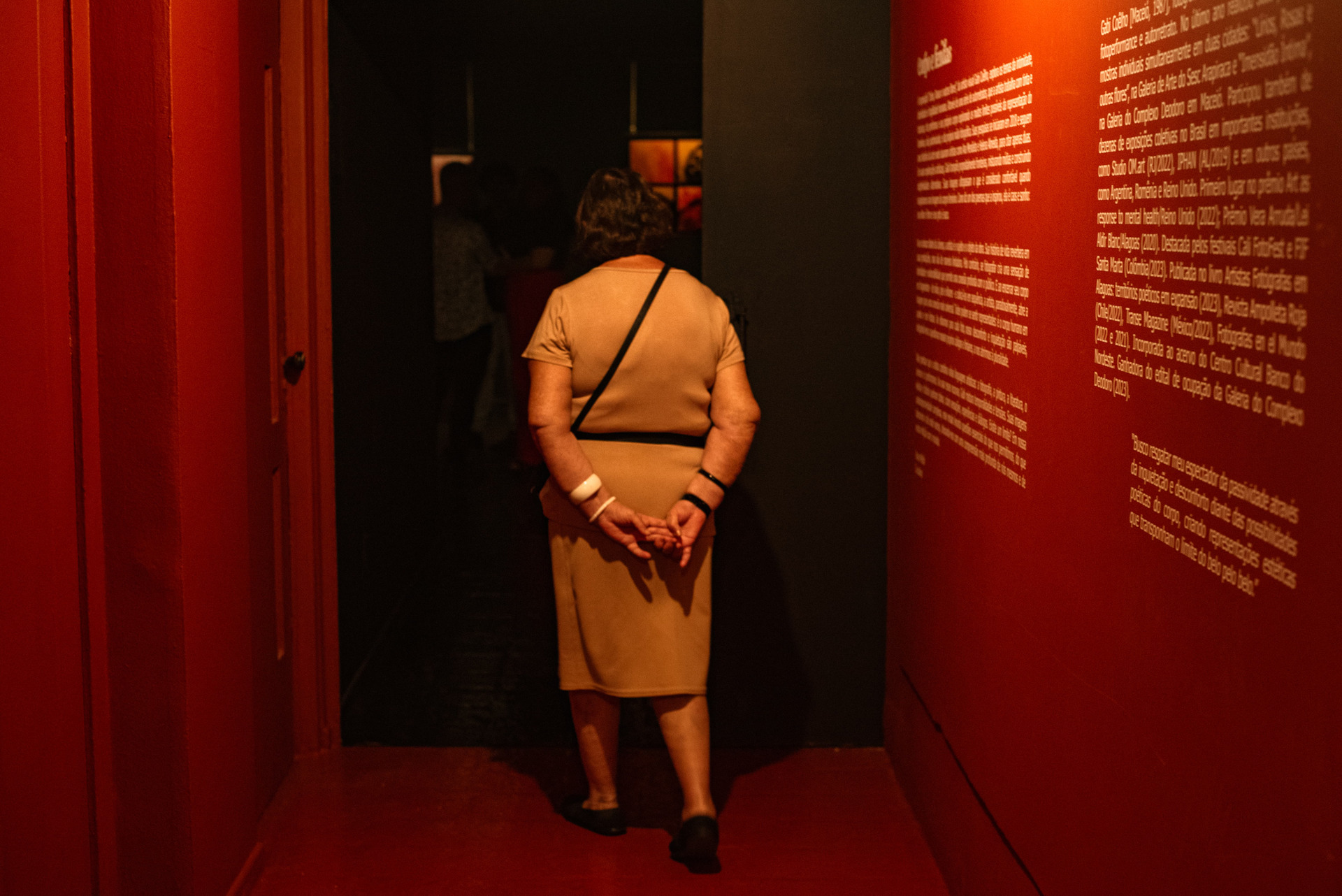 Visitante entrando na galeria da exposição Lírios, Rosas e Outras Flores, de Gabi Coêlho, no Sesc Centro em Maceió, passando pelo texto curatorial em um ambiente escuro e imersivo.